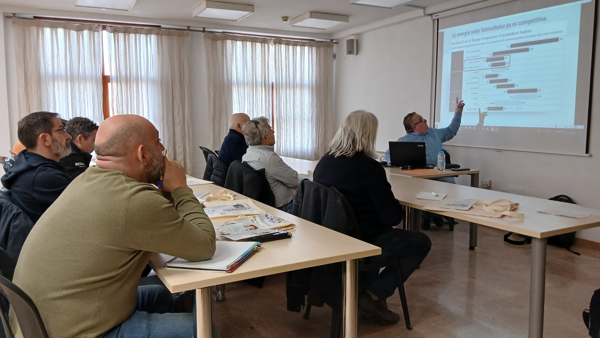 Personas asistentes a una presentación sobre comunidades energéticas en Valderrobres, con un ponente explicando gráficos sobre energía solar fotovoltaica.